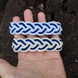 Op de afbeelding: Twee gevlochten armbanden. E&eacute;n is wit met blauwe accenten, de andere is cr&egrave;mekleurig met blauwe accenten. De armbanden zijn gemaakt van dik, geweven touw en worden gepresenteerd op een houten oppervlak.