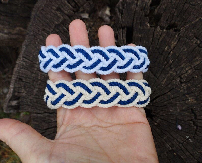 Op de afbeelding: Twee gevlochten armbanden. E&eacute;n is wit met blauwe accenten, de andere is cr&egrave;mekleurig met blauwe accenten. De armbanden zijn gemaakt van dik, geweven touw en worden gepresenteerd op een houten oppervlak.