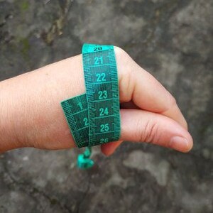 Op de afbeelding: Een groene meetlint om een duim gewikkeld, met metingen in centimeters. De tape heeft witte cijfers op een donkergroene achtergrond. De duim bevindt zich tegen een vage, getextureerde achtergrond.