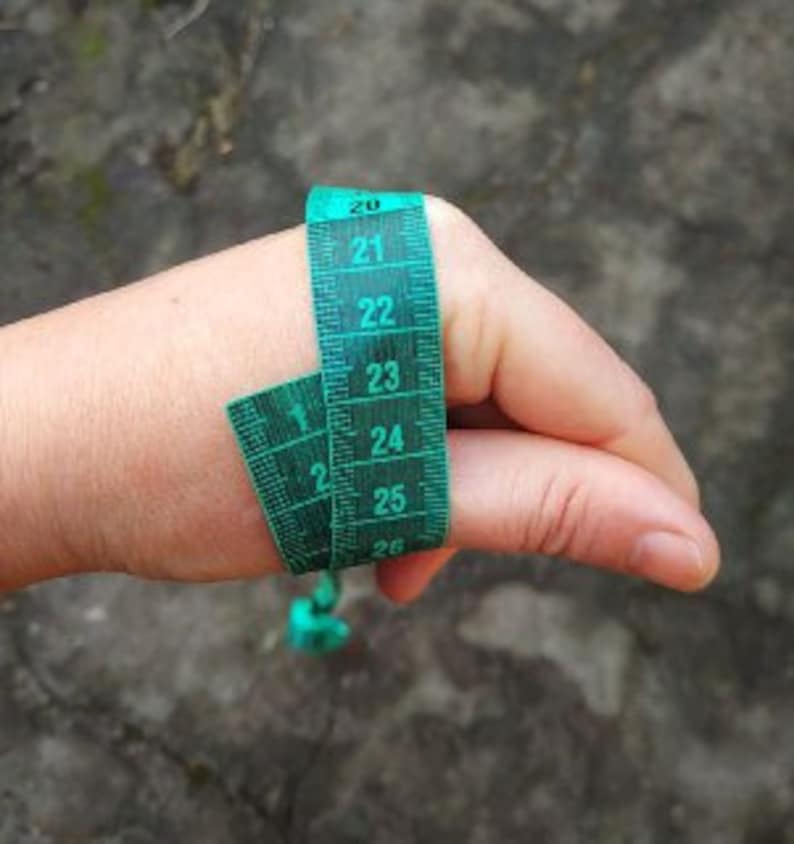Op de afbeelding: Een groene meetlint om een duim gewikkeld, met metingen in centimeters. De tape heeft witte cijfers op een donkergroene achtergrond. De duim bevindt zich tegen een vage, getextureerde achtergrond.