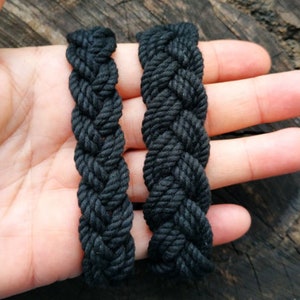 May include: Two black braided rope bracelets, one slightly thicker than the other, are shown on a hand against a wooden background.