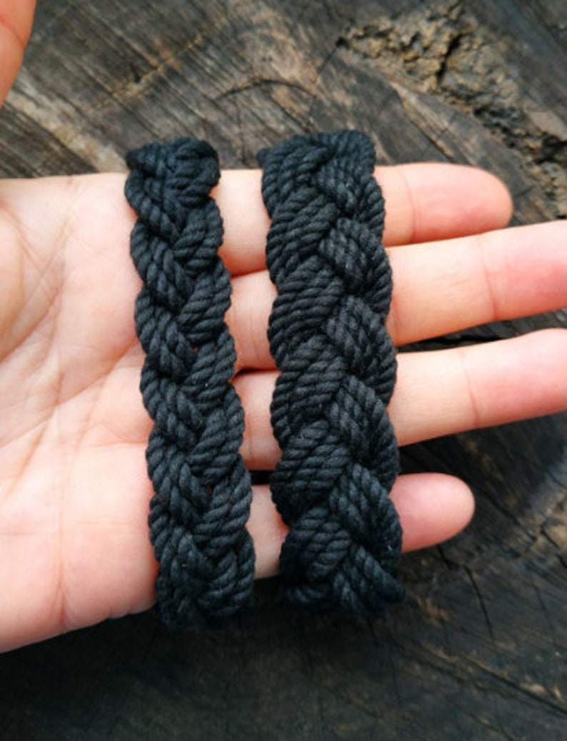 May include: Two black braided rope bracelets, one slightly thicker than the other, are shown on a hand against a wooden background.