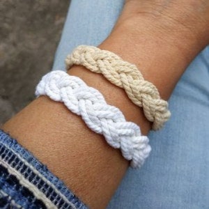 May include: Two braided rope bracelets, one white and one beige, worn on a wrist.