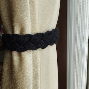 May include: A black braided rope curtain tieback. The tieback is tied around a beige curtain.