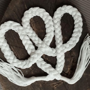 May include: A white braided rope with a tassel end, arranged in a looping pattern. The rope is made of thick, woven material. The background is a dark, textured wooden surface.