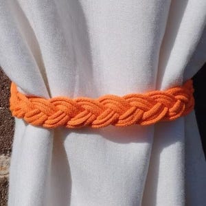May include: An orange braided rope curtain tieback encircles white curtains. The tieback is made of thick, woven rope, creating a textured and decorative element. The bright orange color provides a striking contrast against the white fabric.