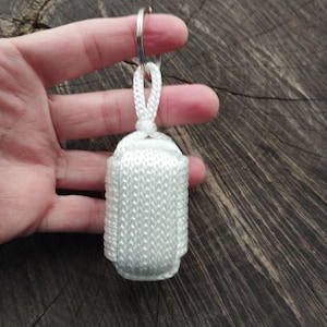 Puede incluir: Un llavero blanco tejido con un anillo plateado. El llavero es rectangular con bordes redondeados y un lazo en la parte superior. El fondo es una superficie de madera oscura y texturizada.