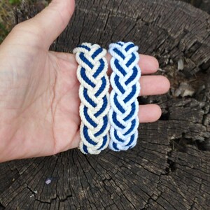 Op de afbeelding: Twee gevlochten armbanden. Elke armband is gemaakt van cr&egrave;mekleurig touw met marineblauwe accenten. De armbanden worden in een hand gehouden tegen een achtergrond van getextureerd, donker hout. De armbanden zijn in nautische stijl.