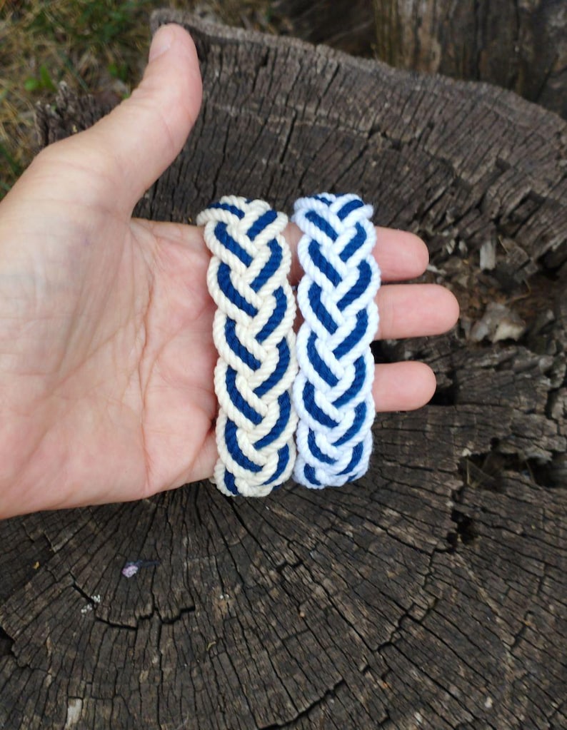 Op de afbeelding: Twee gevlochten armbanden. Elke armband is gemaakt van cr&egrave;mekleurig touw met marineblauwe accenten. De armbanden worden in een hand gehouden tegen een achtergrond van getextureerd, donker hout. De armbanden zijn in nautische stijl.