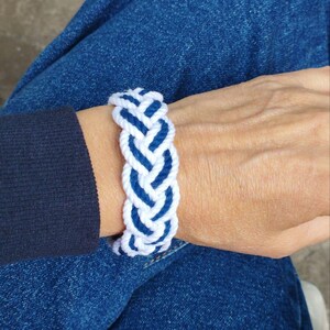 Op de afbeelding: Een gevlochten armband in wit en marineblauw. De armband is gemaakt van dik, geweven touw, met een patroon van afwisselende witte en blauwe secties. De armband wordt om een pols gedragen, met een blauwe spijkerbroek op de achtergrond.