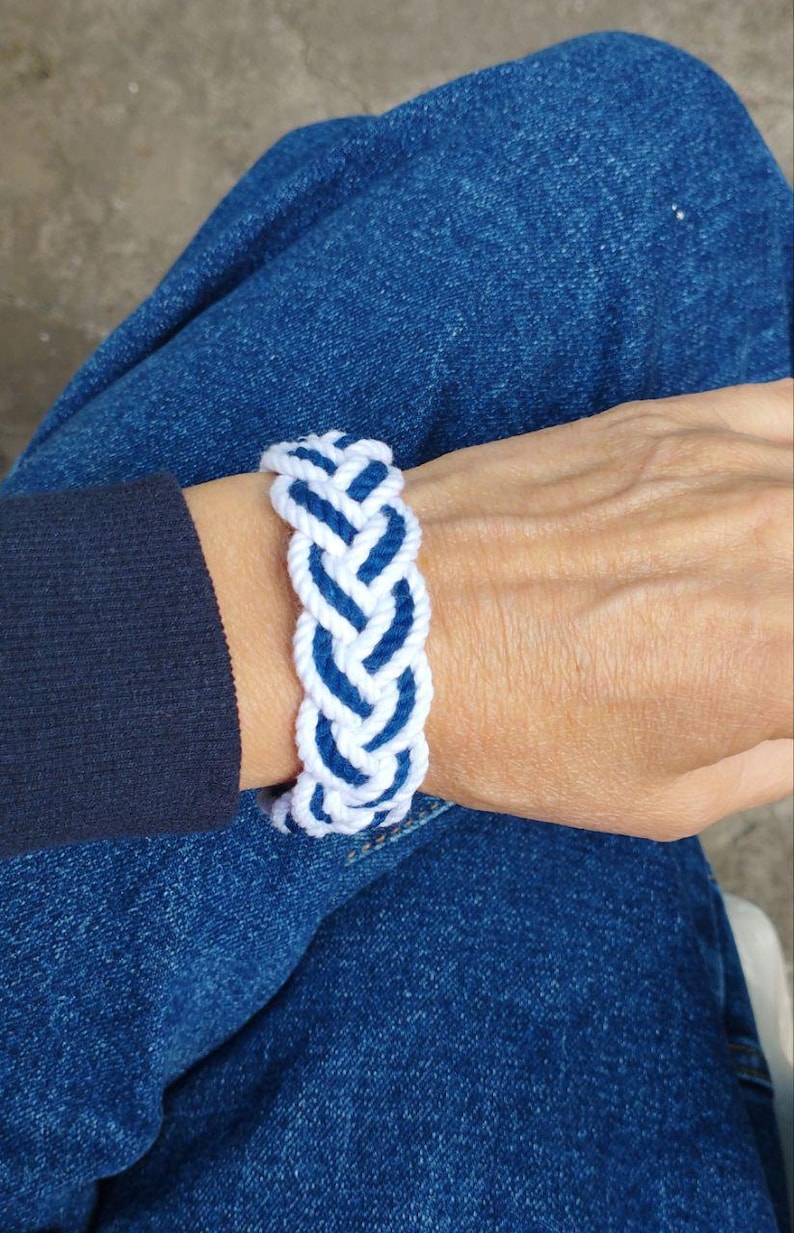 Op de afbeelding: Een gevlochten armband in wit en marineblauw. De armband is gemaakt van dik, geweven touw, met een patroon van afwisselende witte en blauwe secties. De armband wordt om een pols gedragen, met een blauwe spijkerbroek op de achtergrond.