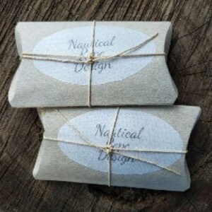 Op de afbeelding: Twee rechthoekige pakketten verpakt in lichtbruin papier, vastgebonden met touw. Elk pakket heeft een wit ovaal etiket met de woorden "Nautical Rose Design" erop. De pakketten zijn gestapeld op een ruw, donkerbruin houten oppervlak.