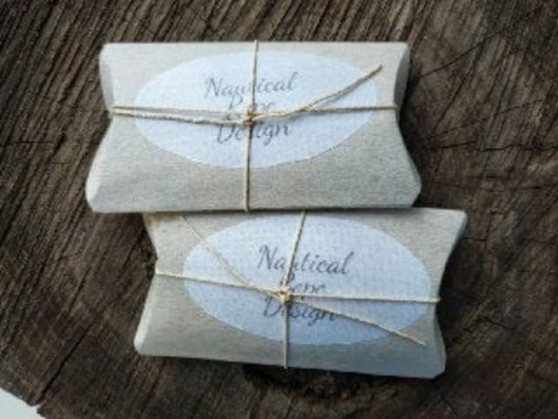 Op de afbeelding: Twee rechthoekige pakketten verpakt in lichtbruin papier, vastgebonden met touw. Elk pakket heeft een wit ovaal etiket met de woorden "Nautical Rose Design" erop. De pakketten zijn gestapeld op een ruw, donkerbruin houten oppervlak.
