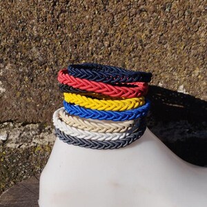 May include: A stack of braided bracelets in various colors, including navy blue, red, yellow, white, and beige. The bracelets are made of a woven material and are stacked on top of each other.