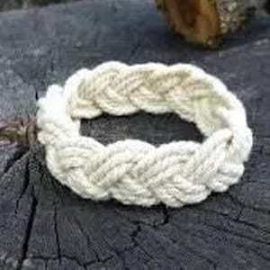 Può includere: Un braccialetto bianco in corda intrecciata, realizzato con un cordoncino spesso e intrecciato. Il braccialetto è appoggiato su una superficie ruvida di colore grigio scuro.