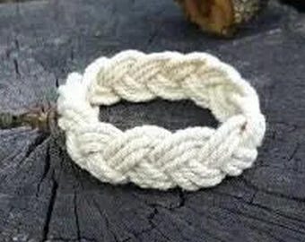 May include: A white braided rope bracelet, made with a thick, woven cord. The bracelet is lying on a rough, dark gray surface.