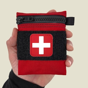 May include: A small red zippered pouch with a black velcro strap and a white cross on a red background. The pouch is designed to hold medical supplies.