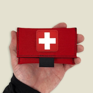 May include: A small red pouch with a white cross on it. The pouch has a black strap and is made of a durable fabric.