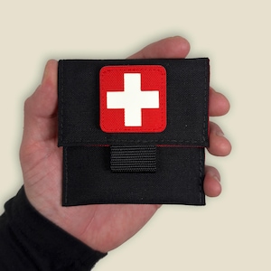 May include: A black first aid pouch with a red and white Swiss cross patch. The pouch has a black strap and is held in a hand.