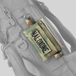 May include: Camouflage-patterned pouch with a zipper and the word "NALOXONE" embroidered on a patch. The pouch is attached to a gray backpack with a metal clip.