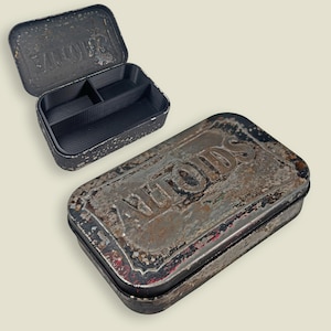 May include: Two vintage Altoids tins, one open and one closed, with a distressed, rusty appearance. The open tin reveals a black interior with dividers. The tins have a rectangular shape and are likely made of metal.