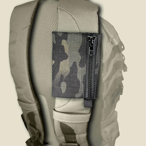 May include: A rectangular, camouflage-patterned pouch with a vertical black zipper closure, attached to the padded shoulder strap of a light tan backpack. The pouch features a dark, multi-tone camouflage print and a black zipper pull tab.