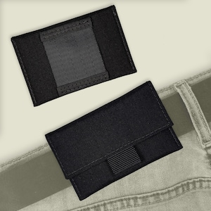 May include: Two black fabric wallets with a velcro closure. The smaller wallet has a single pocket, while the larger wallet has two pockets. The larger wallet is shown on a pair of light gray jeans.