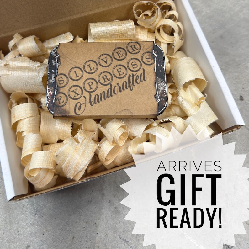 May include: A cardboard box filled with wood shavings, containing a small, rectangular package. The package has a typewriter-style design with the words "SILVER EXPRESS Handcrafted". The image also includes the text "ARRIVES GIFT READY!"