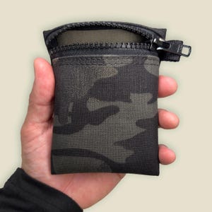 May include: A small, rectangular camouflage pouch with a black zipper and pull tab. The pouch is held open, revealing the interior. The camouflage pattern features shades of green, gray, and black.