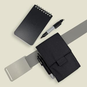 May include: A black spiral-bound notepad, a gray and black marker, and a black fabric pouch attached to a gray belt with a metal buckle. The pouch features an external loop holding a second gray and black marker for quick access.