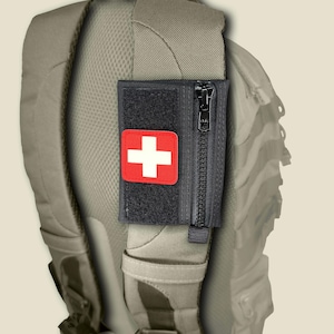 May include: A black rectangular tactical pouch with a vertical zipper closure and a red patch featuring a white cross. The pouch is attached to the padded shoulder strap of a tan backpack, providing a compact storage solution for small items.