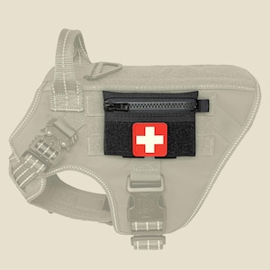 May include: A black rectangular pouch with a zipper closure attached to a light gray dog harness. The pouch features a square red patch with a white cross centered on the front, indicating a medical or first aid kit accessory for the harness.