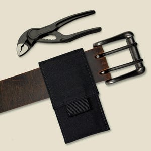 May include: A black multi-tool plier rests above a brown leather belt with a black pouch. The belt has a silver-colored buckle. The plier is made of black metal.