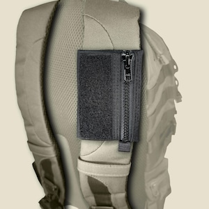 May include: A black rectangular pouch with a vertical zipper and a hook-and-loop fastener panel attached to the beige padded shoulder strap of a backpack. The pouch features a black zipper pull and a textured fabric construction for storage.