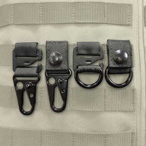 May include: Four black tactical attachment clips and rings secured to a beige MOLLE webbing panel. The set includes two metal carabiner-style hooks and two circular D-rings, each mounted on a black nylon strap with a snap button fastener.