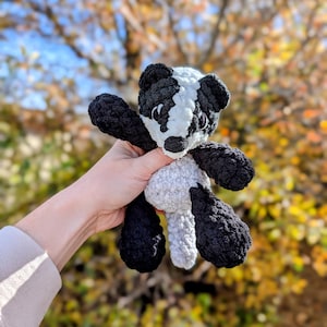 May include: A handmade crochet badger plush toy. The badger has a black and white head, black arms and legs, and a gray body. The plush is held up against a blurred background of fall foliage.
