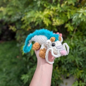 May include: A handmade crochet dragon toy in white, teal, and brown. The dragon has a white body, teal fur accents, and brown paws and horns. The dragon's snout is pink, and it has white whiskers.