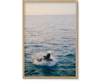 May include: A framed photograph captures a person diving into the ocean, creating a splash. The water is a deep blue, and the sky is a pale yellow. A small sailboat is visible in the distance.