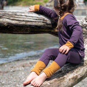 May include: A child's purple long-sleeved top and leggings set with mustard yellow cuffs and trim. The child is sitting on a weathered log near the water. The clothing set is made of a soft material.