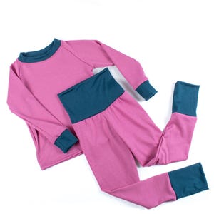 May include: A pink two-piece pyjama set with teal detailing. The long-sleeved top and trousers have teal cuffs, a teal neckline, and a teal waistband. The fabric appears to be soft and lightweight, suitable for sleepwear.