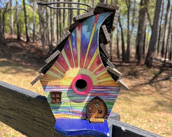 May include: A colorful birdhouse shaped like a wave with a rainbow design and a pink opening. The birdhouse is mounted on a wooden fence.