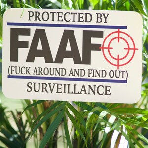 Könnte beinhalten: Ein weißes Schild mit blauem und rotem Text, auf dem "Geschützt durch FAAFO (F**k Around And Find Out) Überwachung" steht. Das Schild hat ein rotes Zielsymbol mit blauem Umriss.