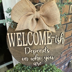 May include: A round wooden sign with a burlap bow and greenery. The sign says "Welcome-ish Depends on who you are".