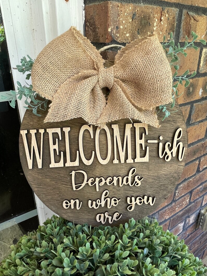 May include: A round wooden sign with a burlap bow and greenery. The sign says "Welcome-ish Depends on who you are".
