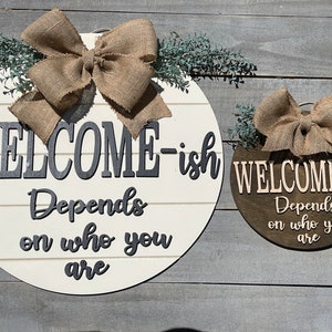 May include: Two round wooden welcome signs with burlap bows and greenery. The larger sign is white with black lettering that reads "WELCOME-ish Depends on who you are". The smaller sign is brown with white lettering that reads "WELCOME-ish Depends on who you are".