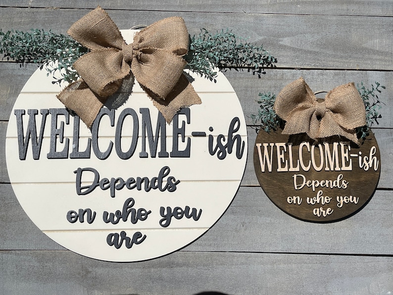 May include: Two round wooden welcome signs with burlap bows and greenery. The larger sign is white with black lettering that reads "WELCOME-ish Depends on who you are". The smaller sign is brown with white lettering that reads "WELCOME-ish Depends on who you are".