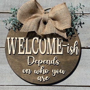 May include: A round wooden sign with a burlap bow and greenery. The sign says "Welcome-ish Depends on who you are".