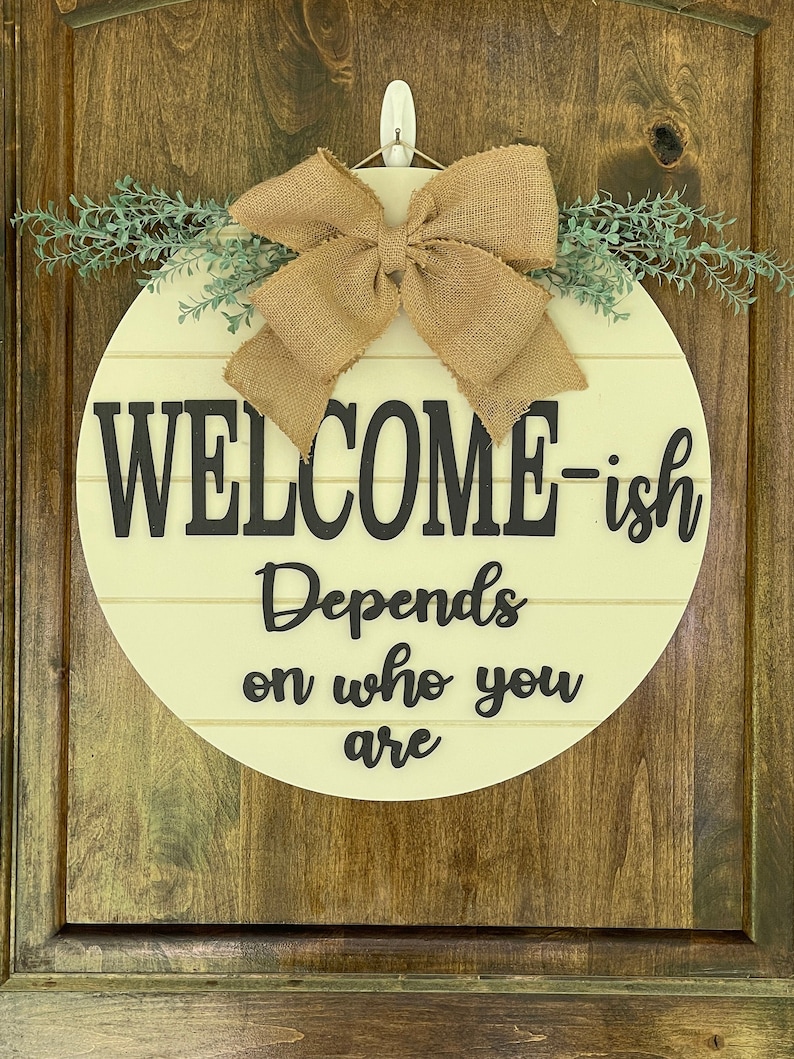 May include: A round wooden sign with a burlap bow and greenery. The sign says "Welcome-ish Depends on who you are".