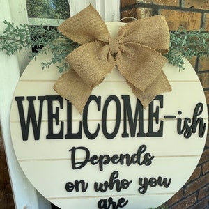 May include: A round wooden door hanger with a burlap bow and greenery. The sign reads "Welcome-ish Depends on who you are".