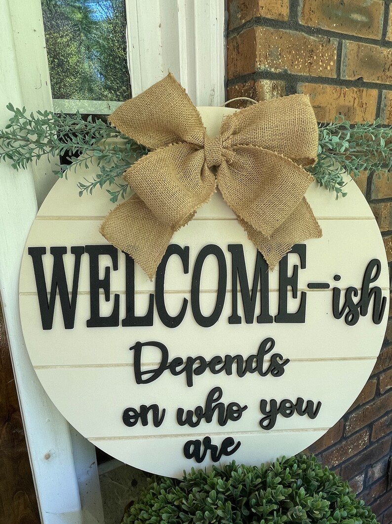 May include: A round wooden door hanger with a burlap bow and greenery. The sign reads "Welcome-ish Depends on who you are".